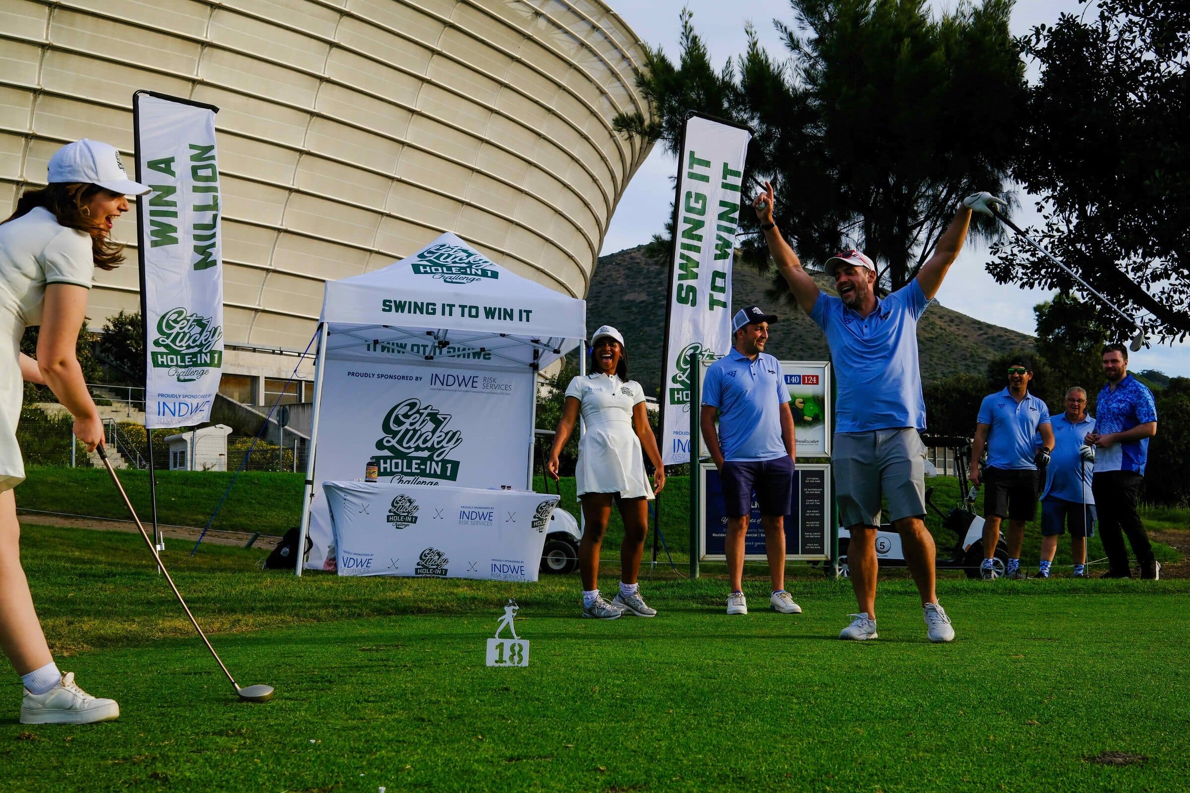 A golfer celebrating with both arms up on the Get Lucky challenge tee, beside the Swing It To Win It gazebo